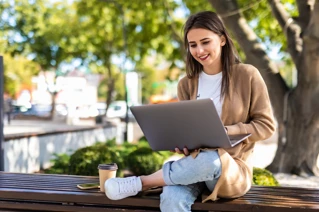 En kvinna som jobbar utomhus med en laptop på en bänk under ett träd